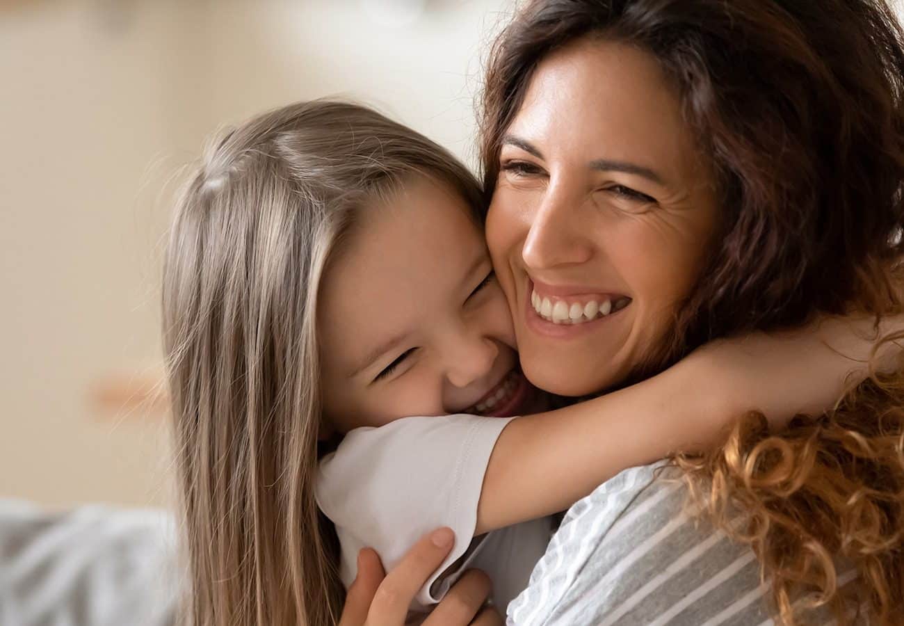 A young girl and a woman share a joyful embrace, both smiling with their eyes closed, conveying warmth and happiness in a close-up portrait.