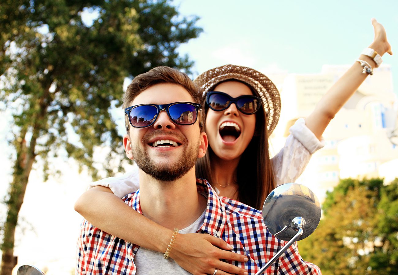 A smiling man and woman wearing sunglasses and casual clothes ride a scooter outdoors. The woman has her arms around the man and raises one arm, both appearing joyful. Trees and buildings are visible in the background.