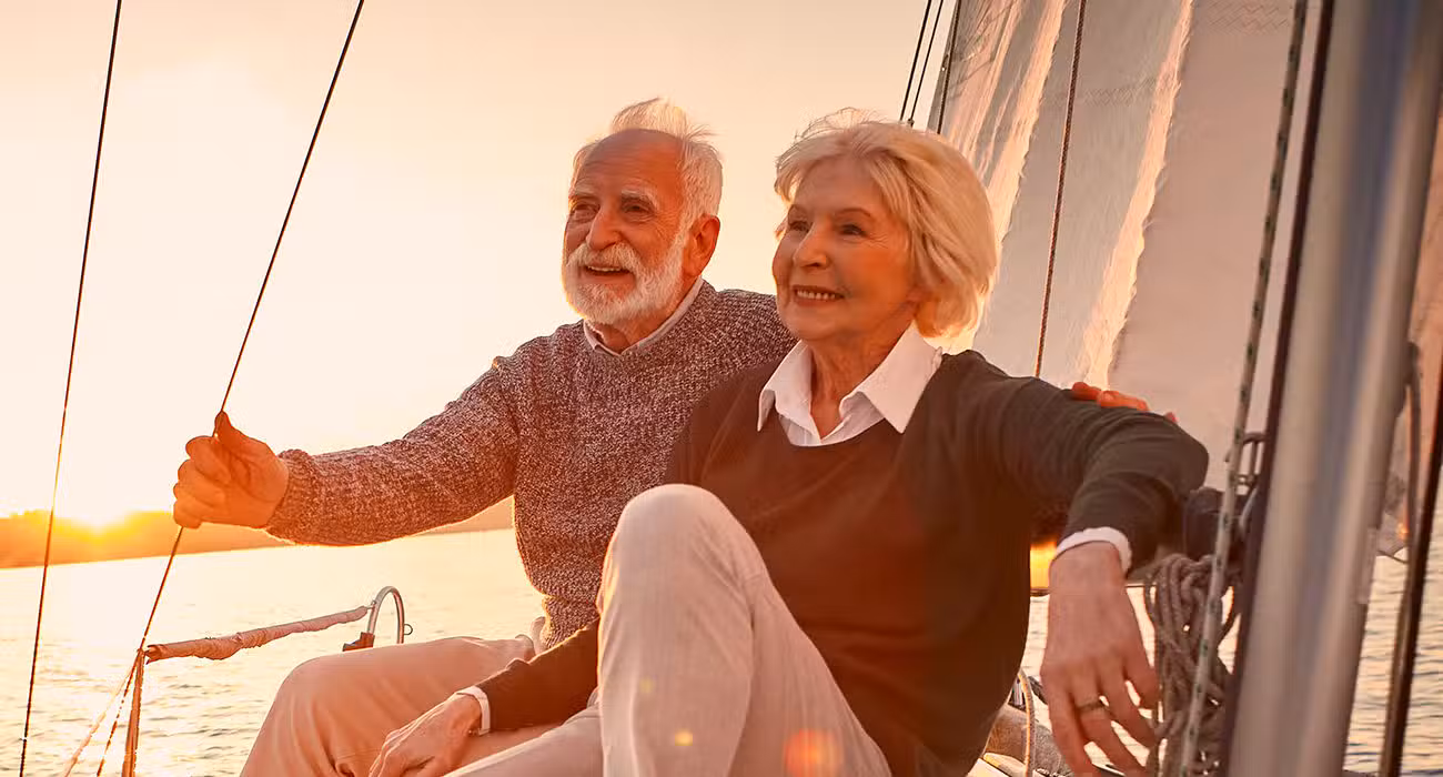 An elderly couple sits together on a sailboat at sunset, smiling and relaxed, with warm sunlight illuminating their faces and the sail in the background.