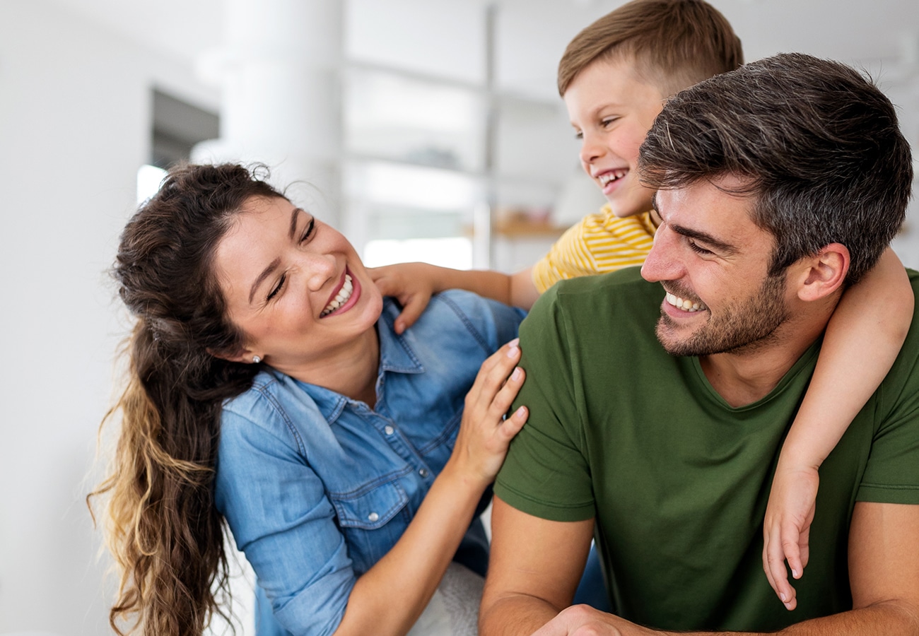 A woman, a man, and a young boy smile and laugh together indoors. The boy, wearing a yellow shirt, hugs the mans shoulders from behind as the woman, in a blue shirt, looks at them happily.