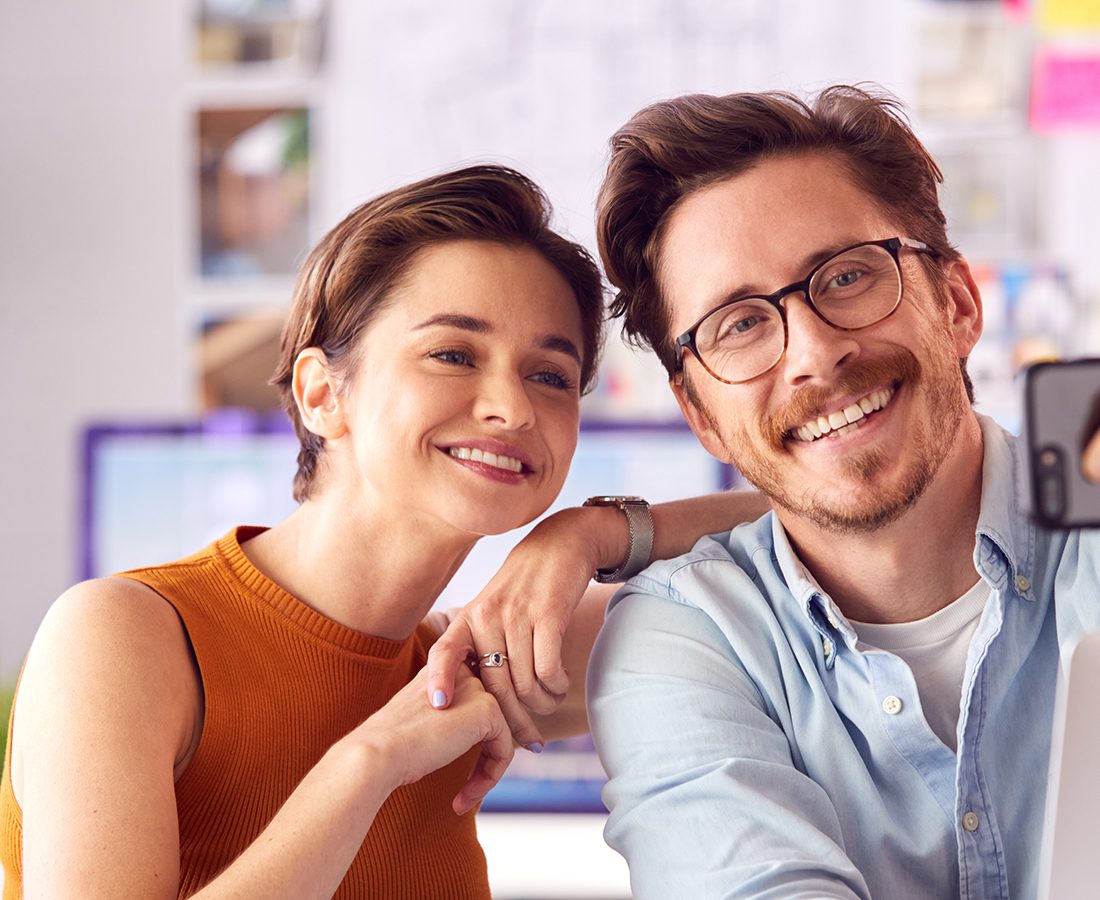 A smiling woman in an orange sleeveless top and a man with glasses in a light blue shirt pose closely together, taking a selfie indoors.
