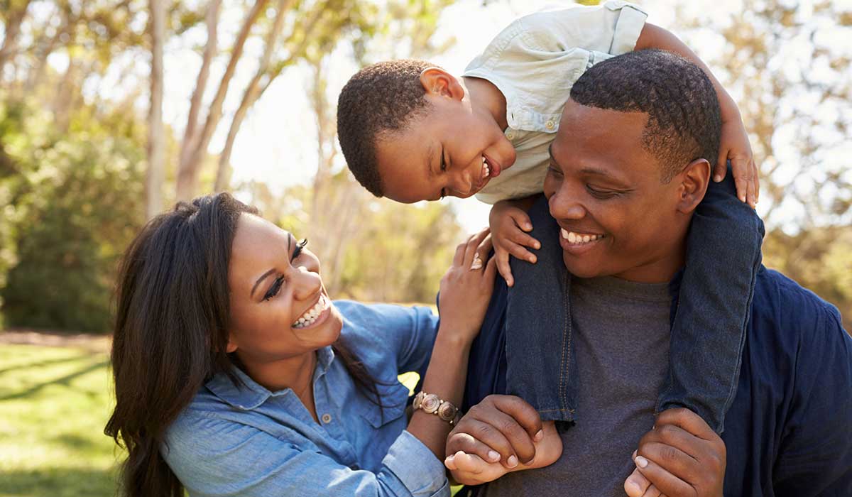 A smiling couple stands outdoors, with the man carrying a laughing young boy on his shoulders. The woman touches the mans arm. Sunlight filters through the trees in the background.
